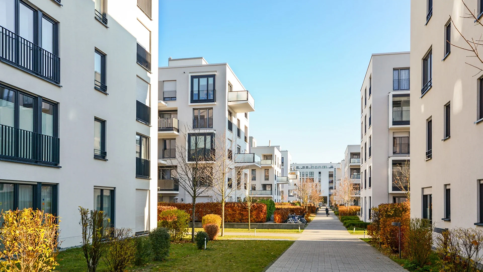 Moderne Apartmentgebäude in einer grünen Wohngegend innerhalb einer Stadt. Die Architektur der Gebäude ist zeitgemäß, mit großen Fenstern und klaren Linien, die sich harmonisch in die natürliche Umgebung mit viel Grün und Bäumen einfügen.