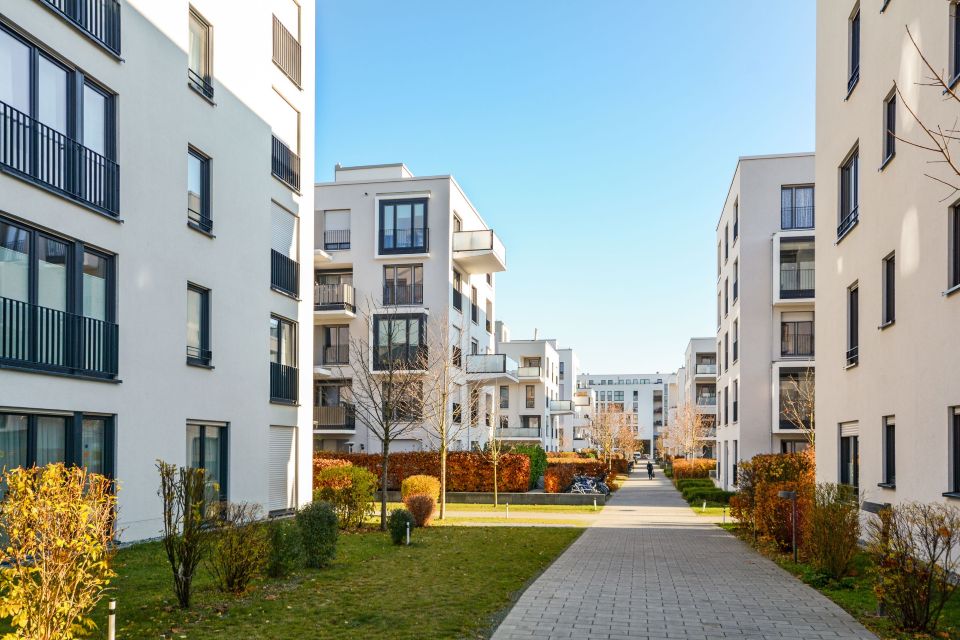 Moderne Apartmentgebäude in einer grünen Wohngegend innerhalb einer Stadt. Die Architektur der Gebäude ist zeitgemäß, mit großen Fenstern und klaren Linien, die sich harmonisch in die natürliche Umgebung mit viel Grün und Bäumen einfügen.